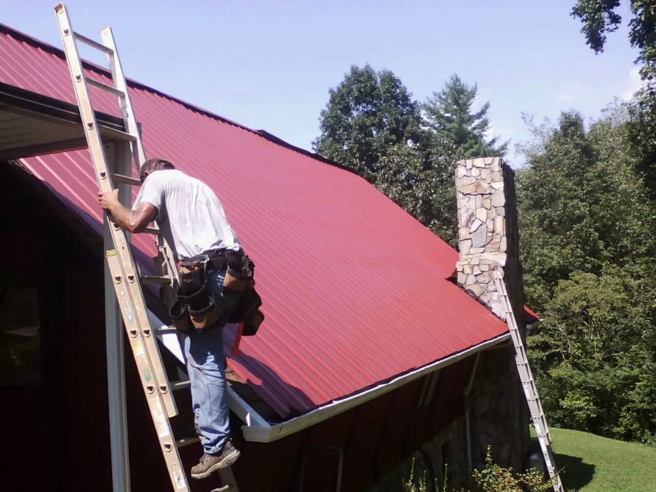 Roofer on a ladder beside a red metal roof and stone chimney — Soffit & Fascia Repair by Roofing Brothers in Pleasant Plain